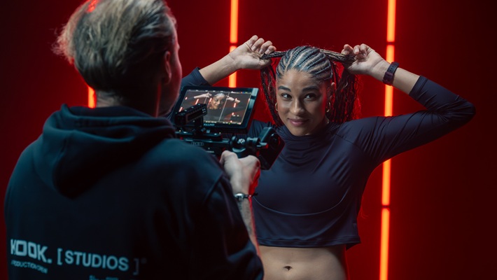 Die Fussballerin Sydney Schertenleib mit geflochtenem Haar posiert vor einer Kamera in einem Studio mit rotem Hintergrund. Es handelt sich dabei um eine behind the scenes-Aufnahme für eine Werbefilmproduktion für die Uhrenmarke Norqain produziert im Mietstudio für Film- und Fotoproduktionen StudioGutenswil in der Nähe von Zürich (Schweiz).