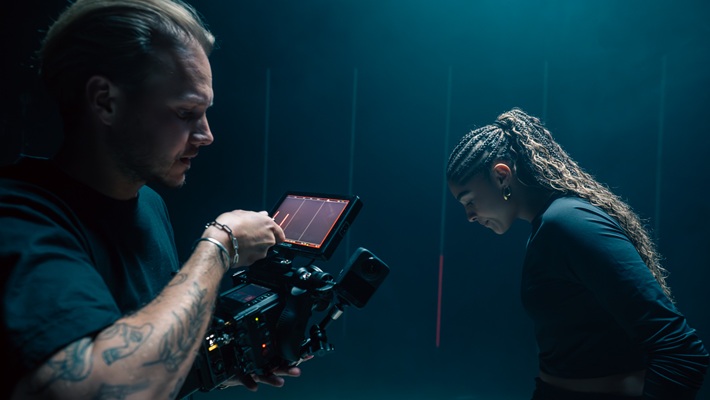 Ein Mann bedient eine Kamera, während die Fussballerin Sydney Schertenleib nachdenklich nach unten schaut. Es handelt sich dabei um eine behind the scenes-Aufnahme für eine Werbefilmproduktion für die Uhrenmarke Norqain produziert im Mietstudio für Film- und Fotoproduktionen StudioGutenswil in der Nähe von Zürich (Schweiz).